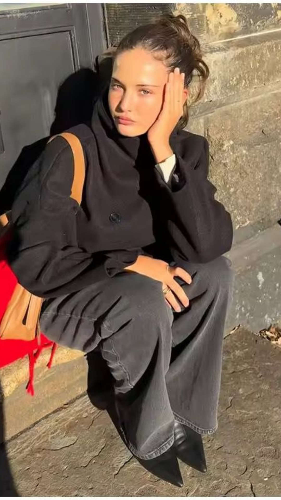 Woman sitting on a step wearing a black coat and gray pants, with a red bag beside her.