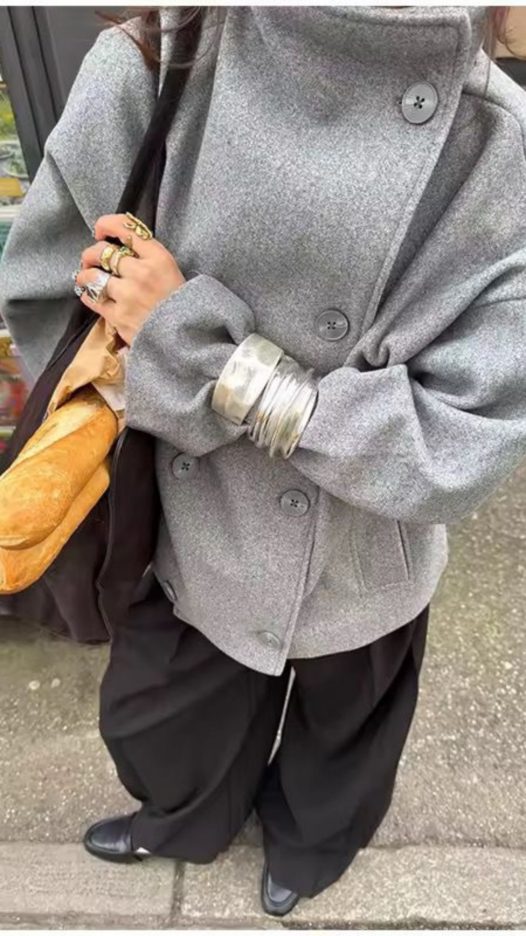Person wearing a gray coat holding a baguette and a metallic cup on a sidewalk.